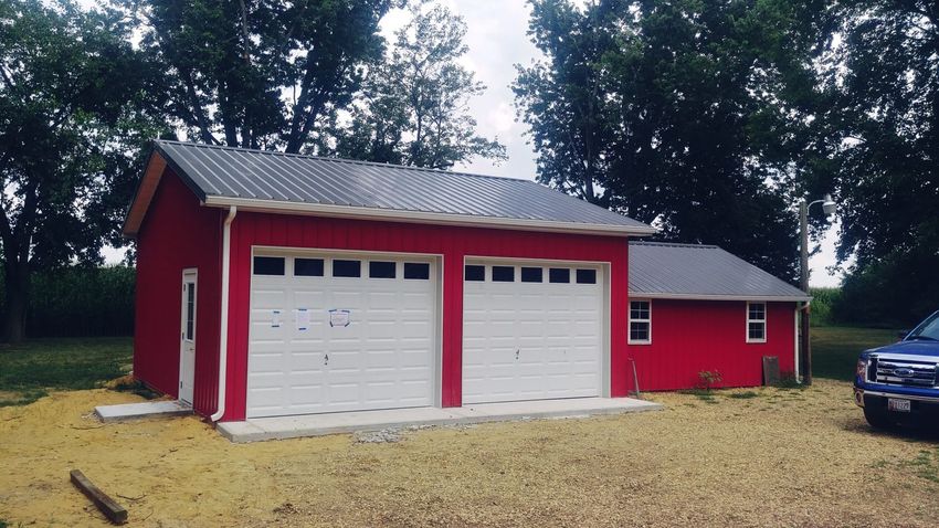 Red garage with two white doors and gray roof beside a small red shed, on a gravel driveway.