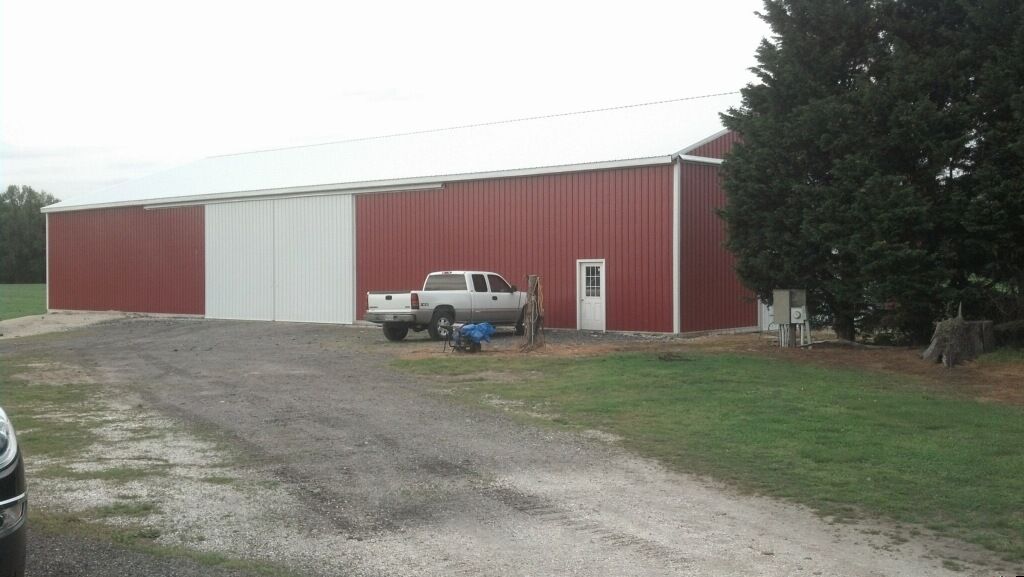 Red metal barn with white doors beside a gravel driveway, with a pickup truck parked out front.