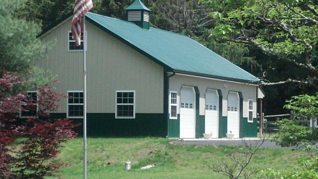 Green barn-like building with a teal roof, white garage doors, and an American flag on a grassy lot