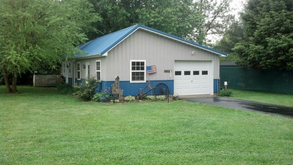 Small beige house with blue trim and garage, surrounded by a green lawn and trees.