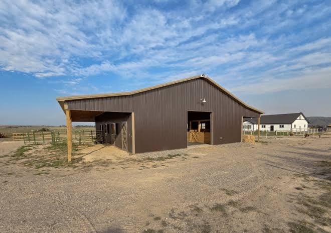 Brown barn on a dusty rural lot under a cloudy blue sky, with a white house in the background.