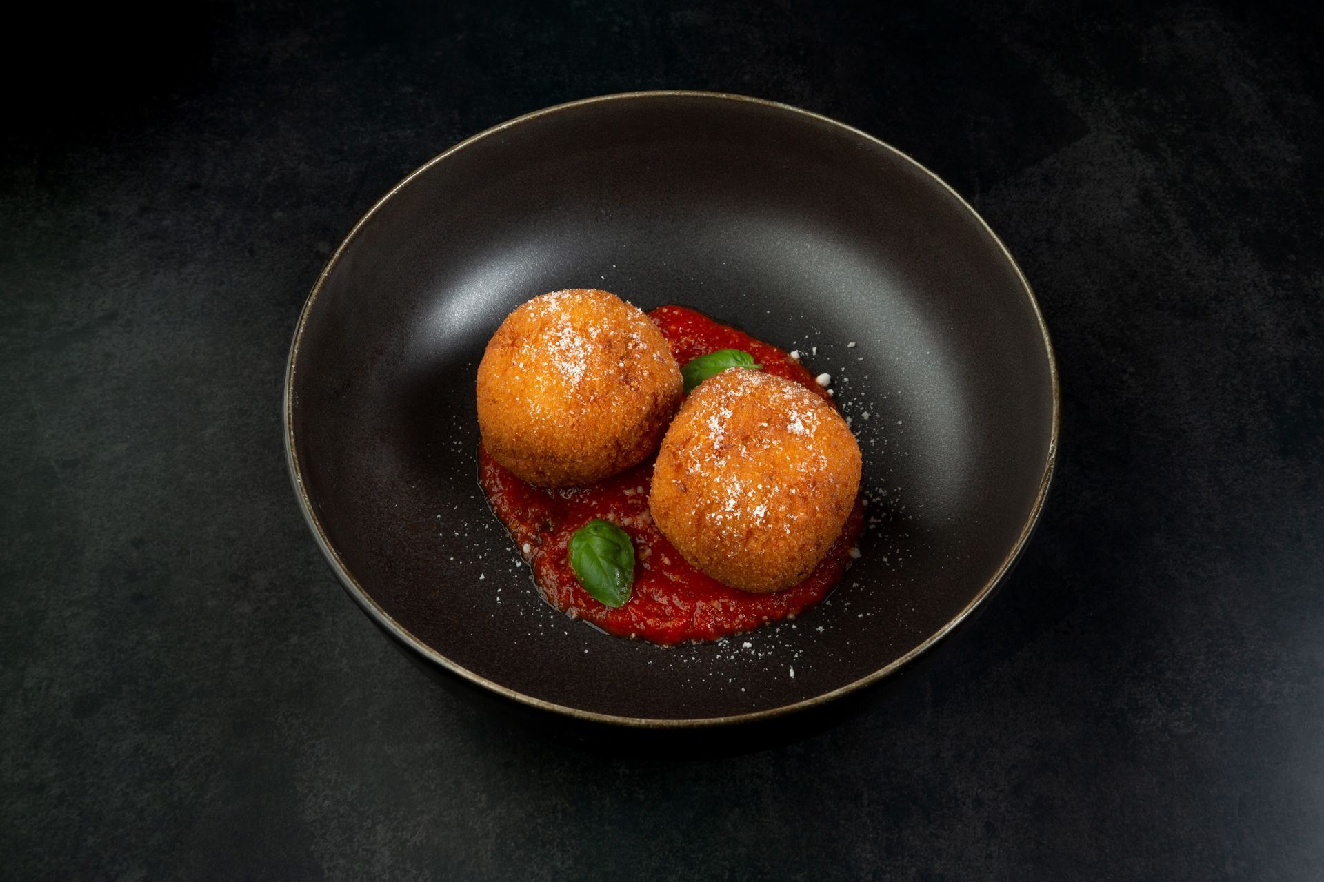 Deux croquettes frites à la sauce tomate, servies sur une assiette sombre avec des feuilles de basilic.