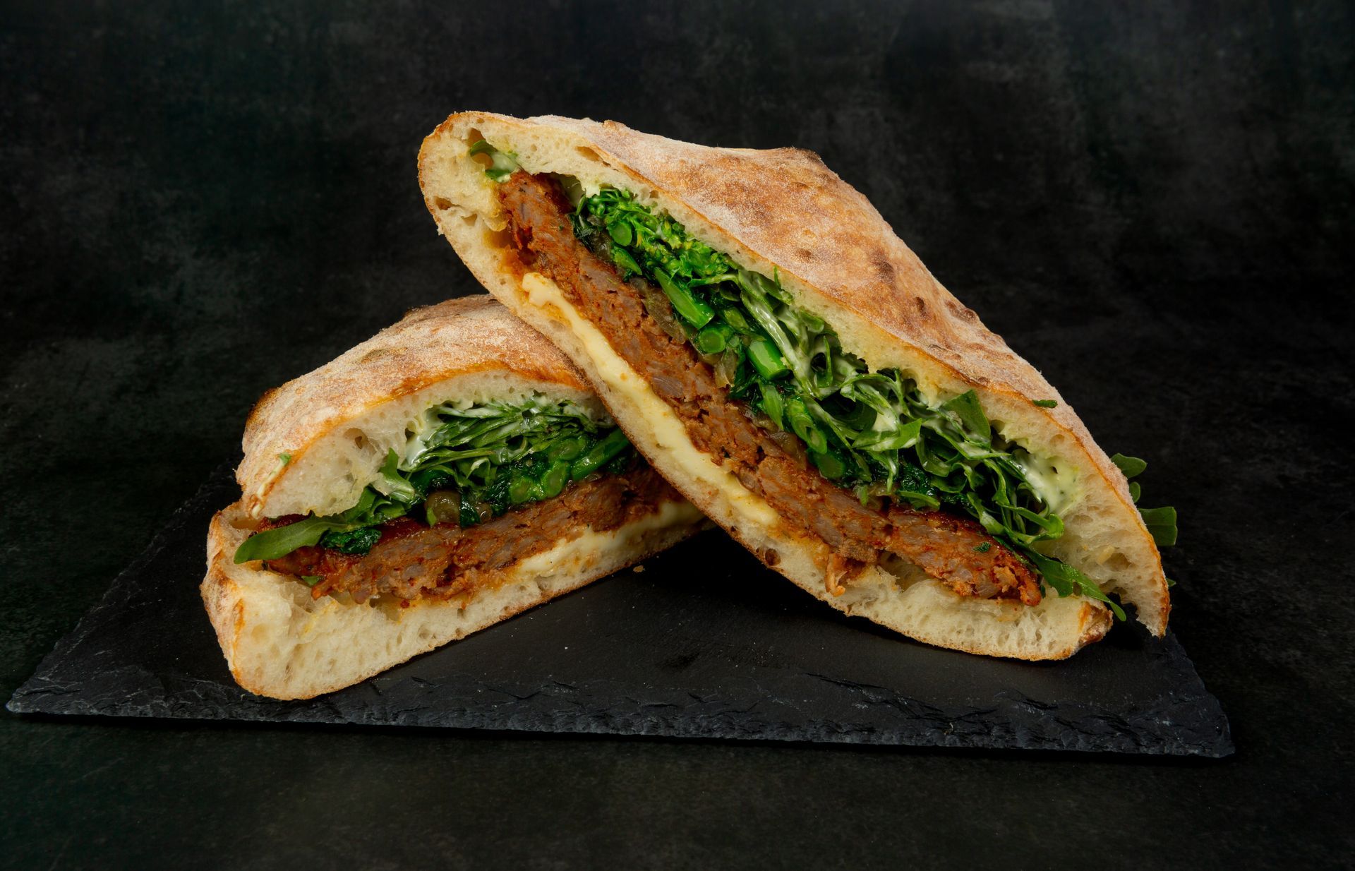 Deux moitiés de sandwich grillées garnies de salade verte, servies sur une planche d'ardoise sombre.
