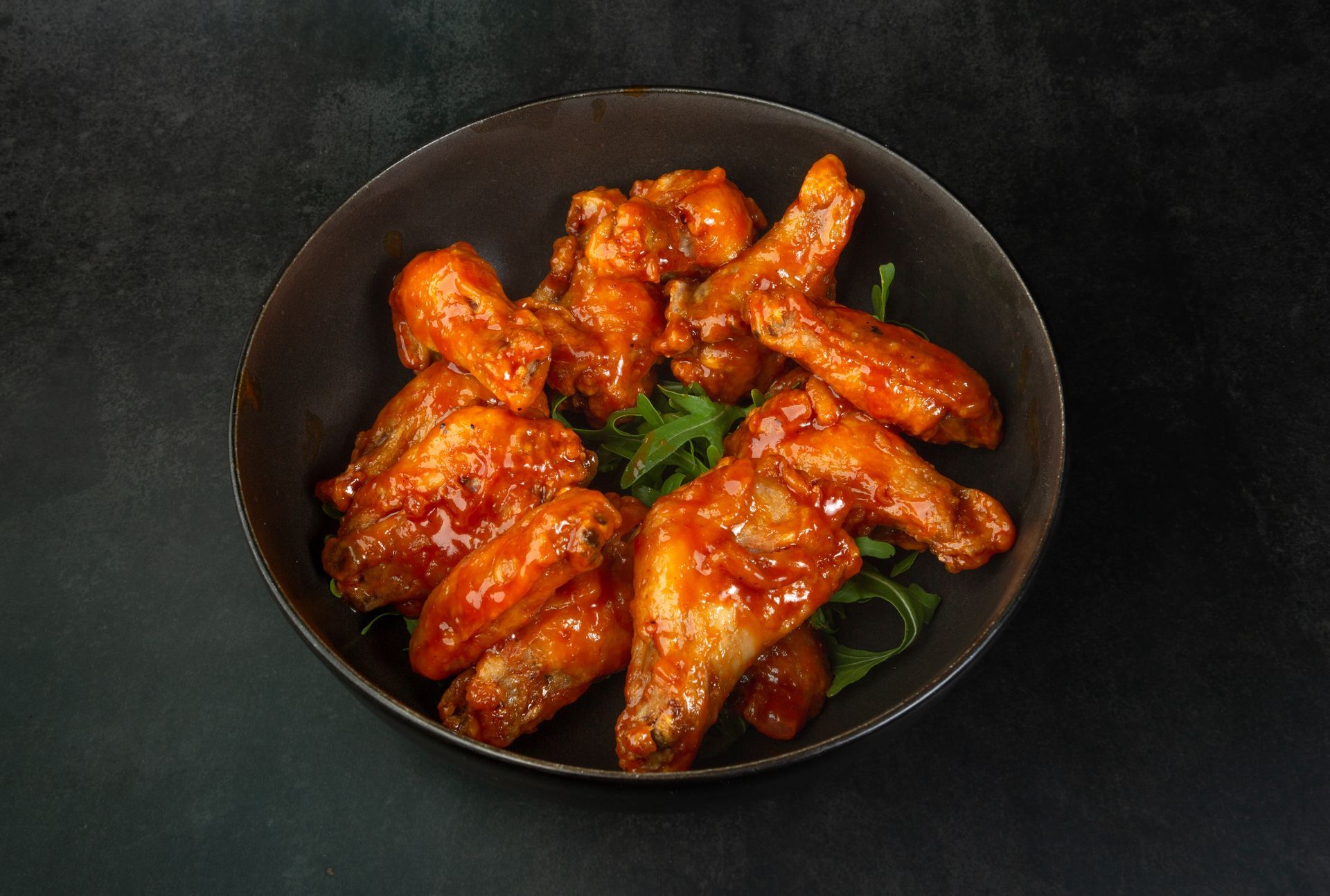 Ailes de poulet épicées et glacées dans un bol noir, garnies d'herbes sur fond sombre.