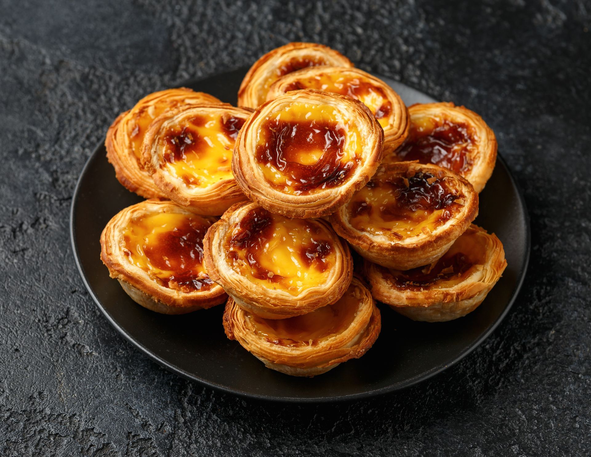 Assiette de tartelettes à la crème dorées empilées sur un plat noir, sur fond sombre.