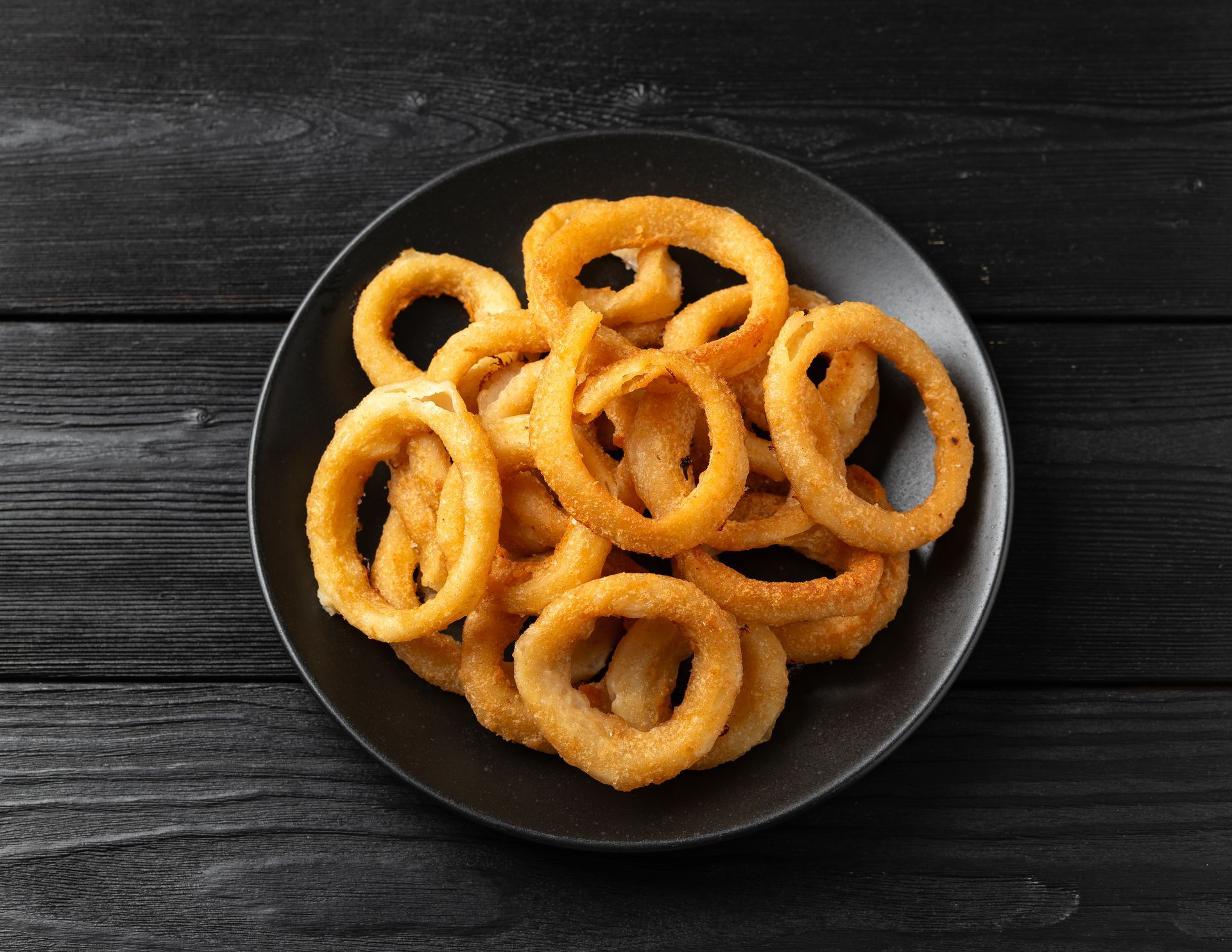 Rondelles d'oignon frites sur une assiette noire, posée sur une table en bois sombre