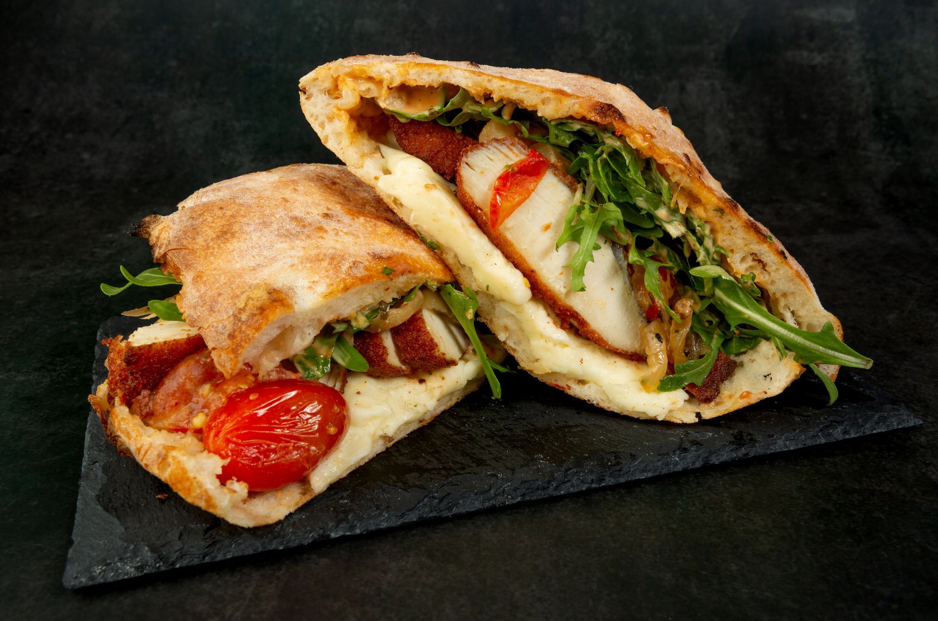 Sandwichs pita farcis et coupés en tranches, avec tomates, salade et poulet grillé, servis sur une planche d'ardoise foncée.