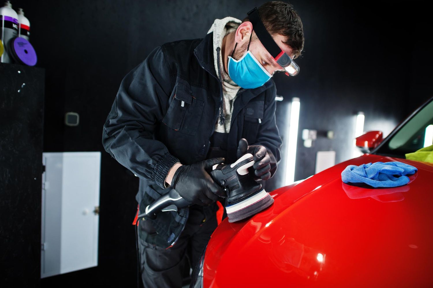 A technician wearing a mask and headlamp uses a rotary buffer to polish the hood of a red car in a workshop.