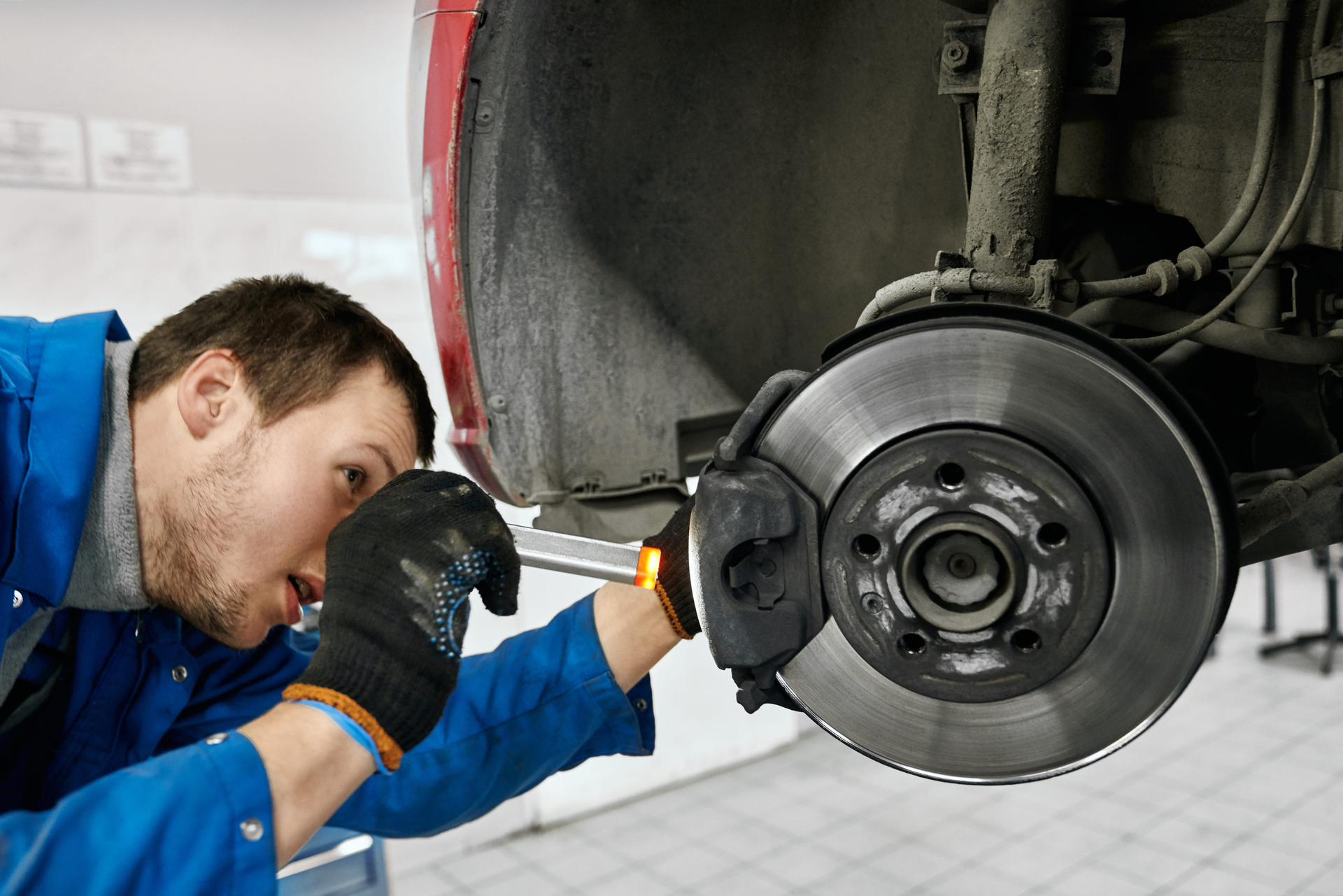 A mechanic in a blue uniform and black gloves inspects a vehicle's brake disc and caliper in a repair shop.