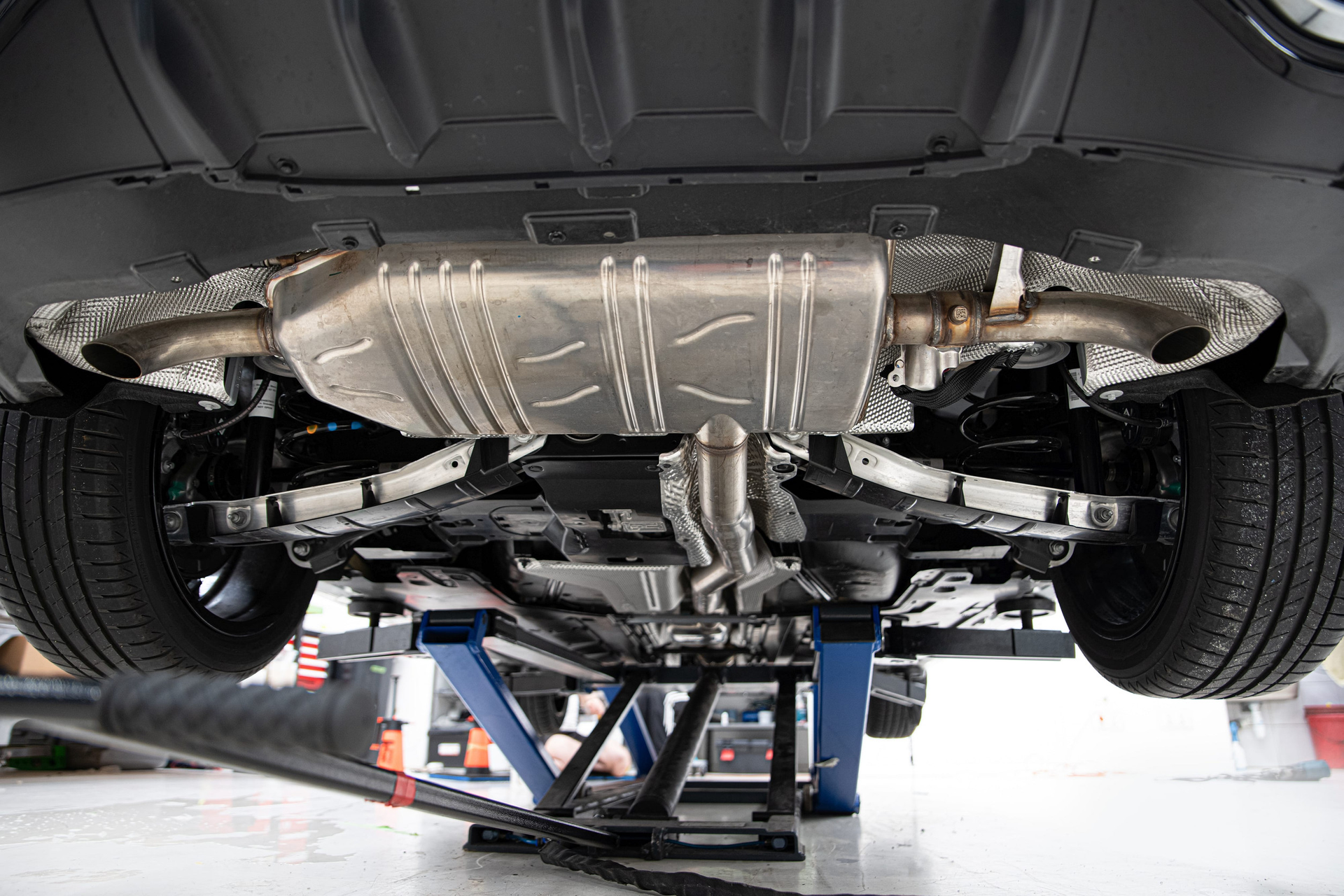 A low-angle view of a car’s undercarriage, showing the exhaust system, tires, and suspension on a hydraulic lift.