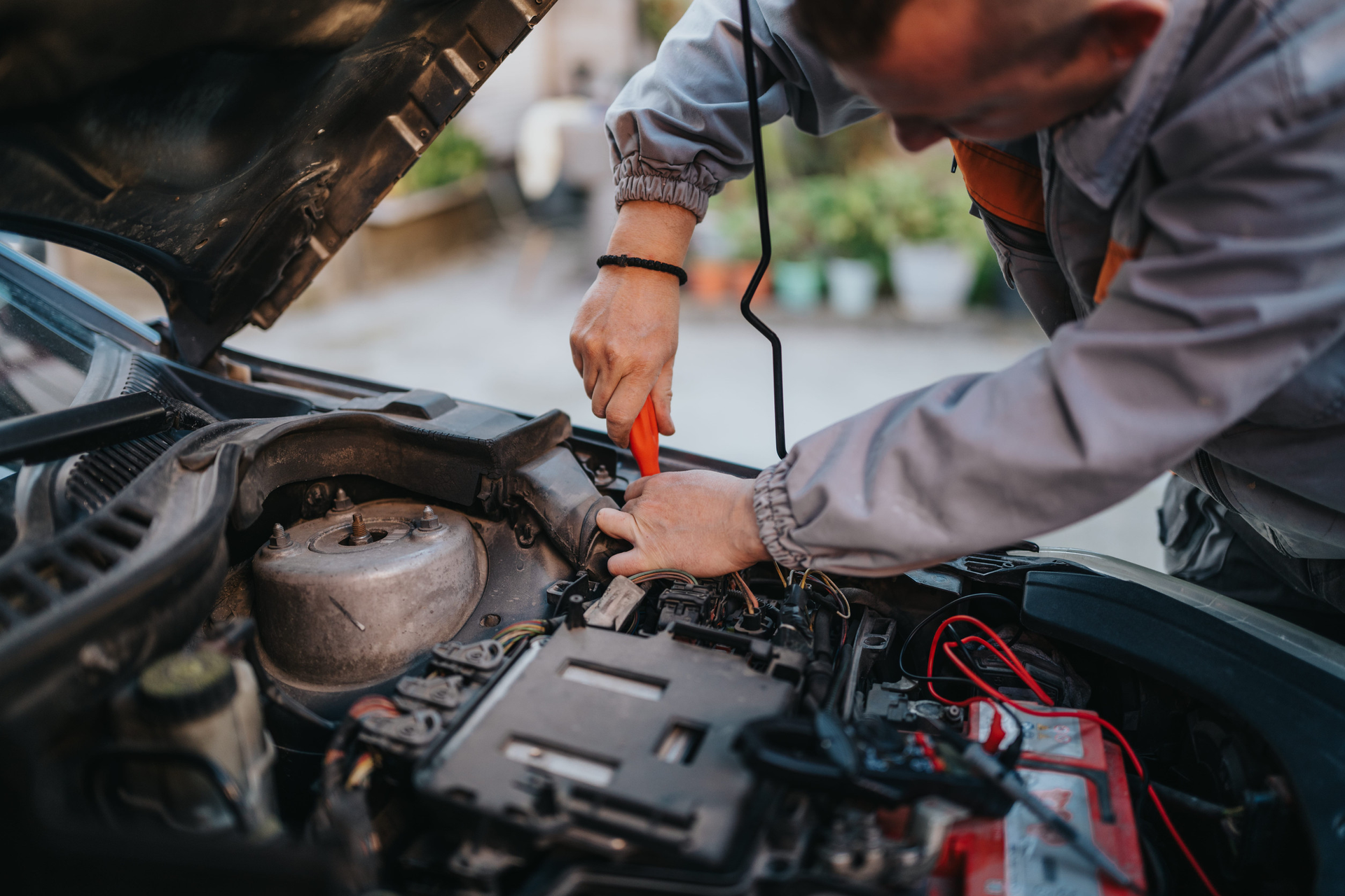 A mechanic in a grey uniform works on an open car engine, using a red-handled tool to make adjustments.