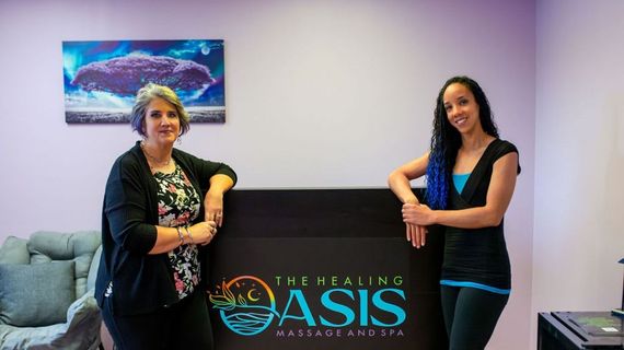 Two women stand beside a table with a “Healing Oasis” sign in a lobby, smiling at the camera.