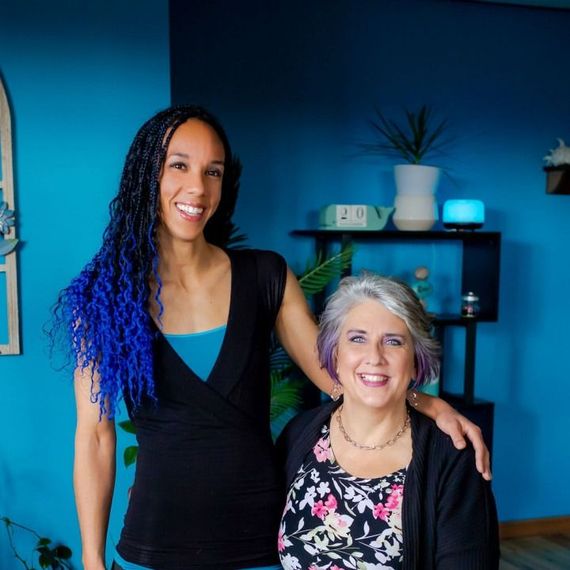 Two smiling women standing together in a blue room with a shelf and plant behind them