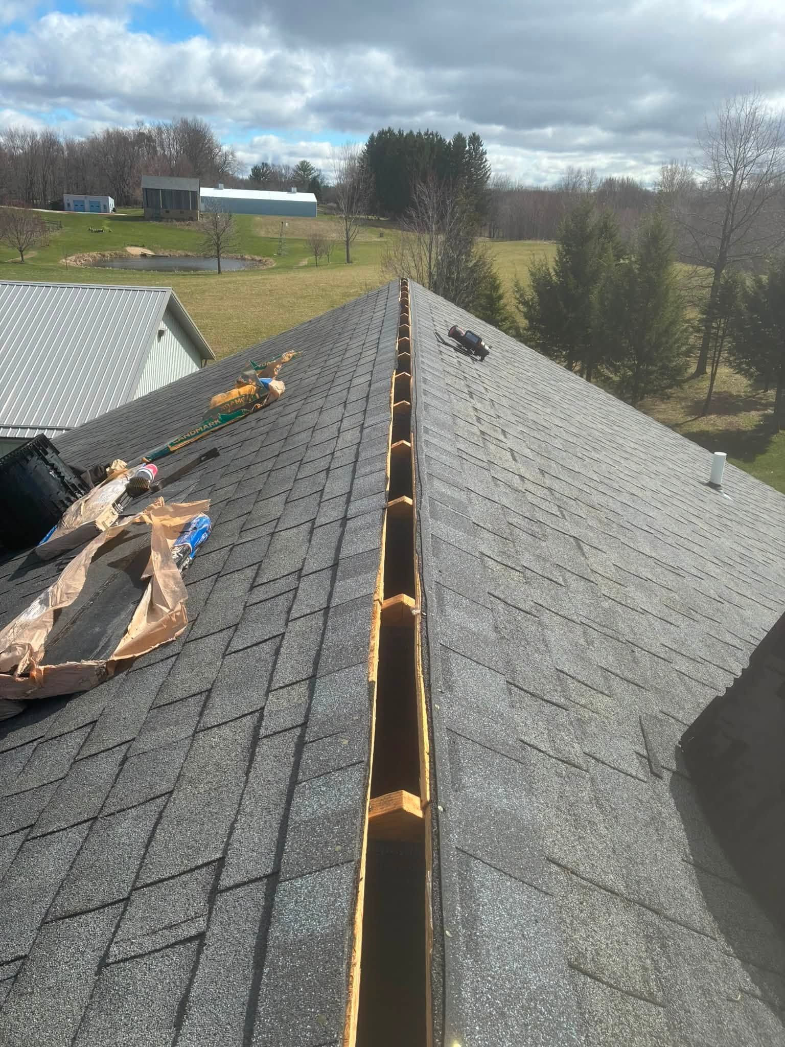 Roof under repair with exposed ridge line and tools on gray shingles, overlooking a rural yard