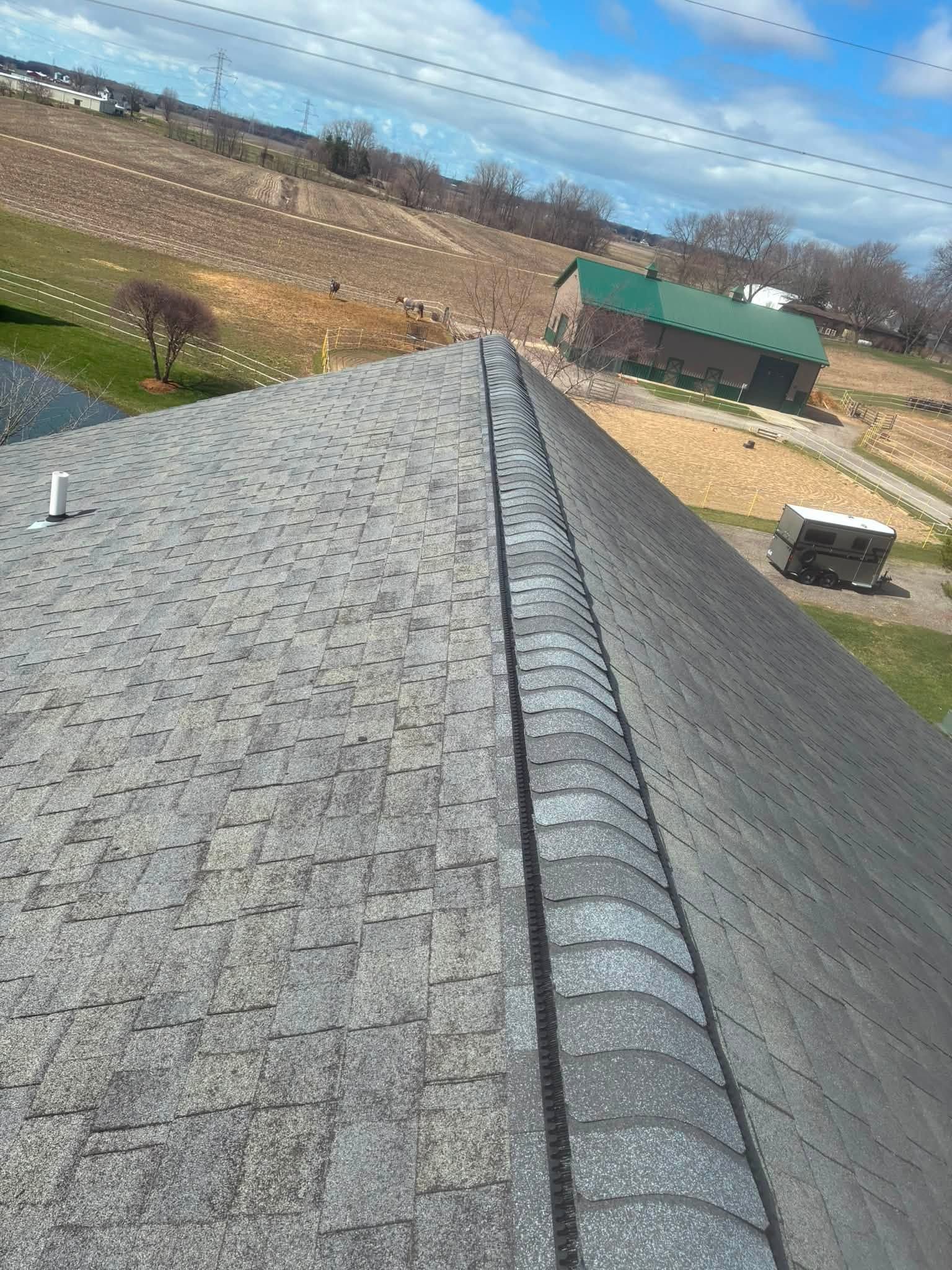 View from a shingled roof over rural fields, a green barn, and a partly cloudy sky