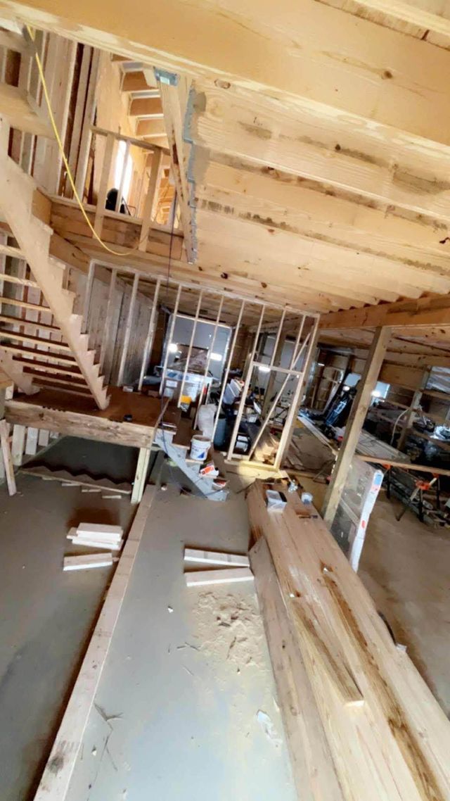 Inside a wood-framed building under construction, stacked lumber and workers are visible through unfinished walls.