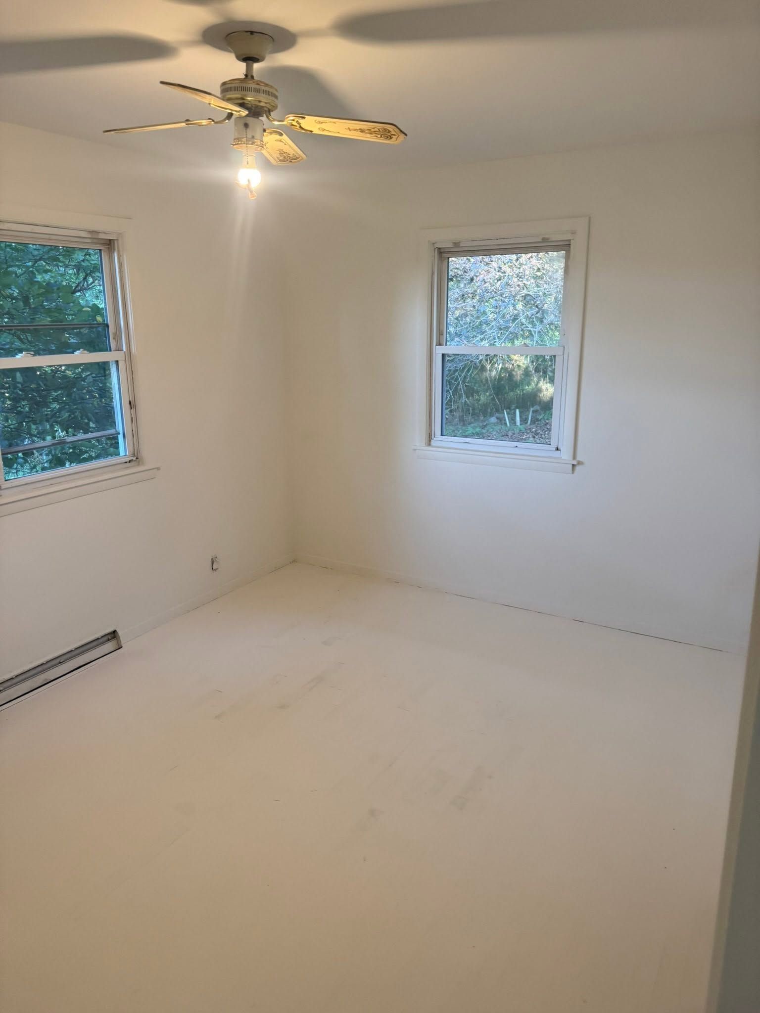 Empty white bedroom with two windows, ceiling fan, and baseboard heater