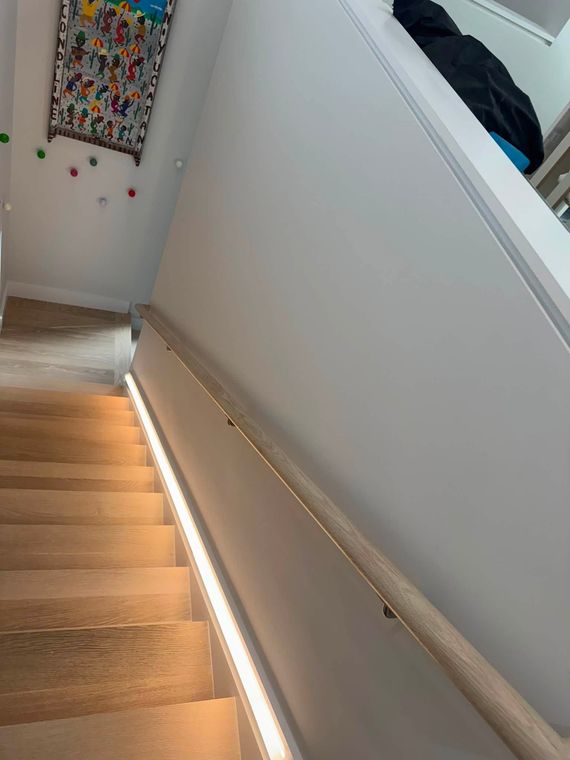 Stairwell with wooden steps, white walls, and a handrail lit by a strip light.