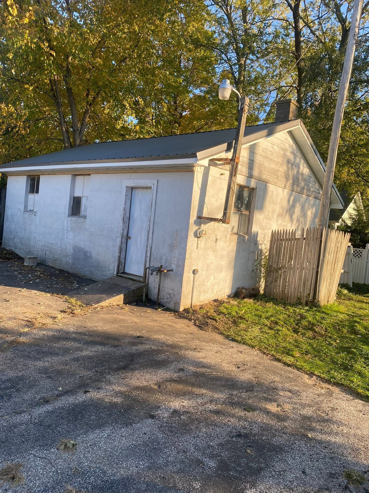 Small white outbuilding with a slanted roof and peeling paint beside a gravel driveway and trees.