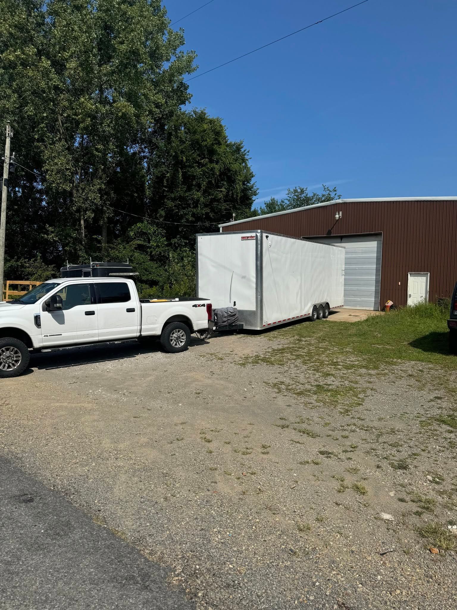 White pickup truck parked beside a white box trailer and brown building on a gravel lot under a clear blue sky