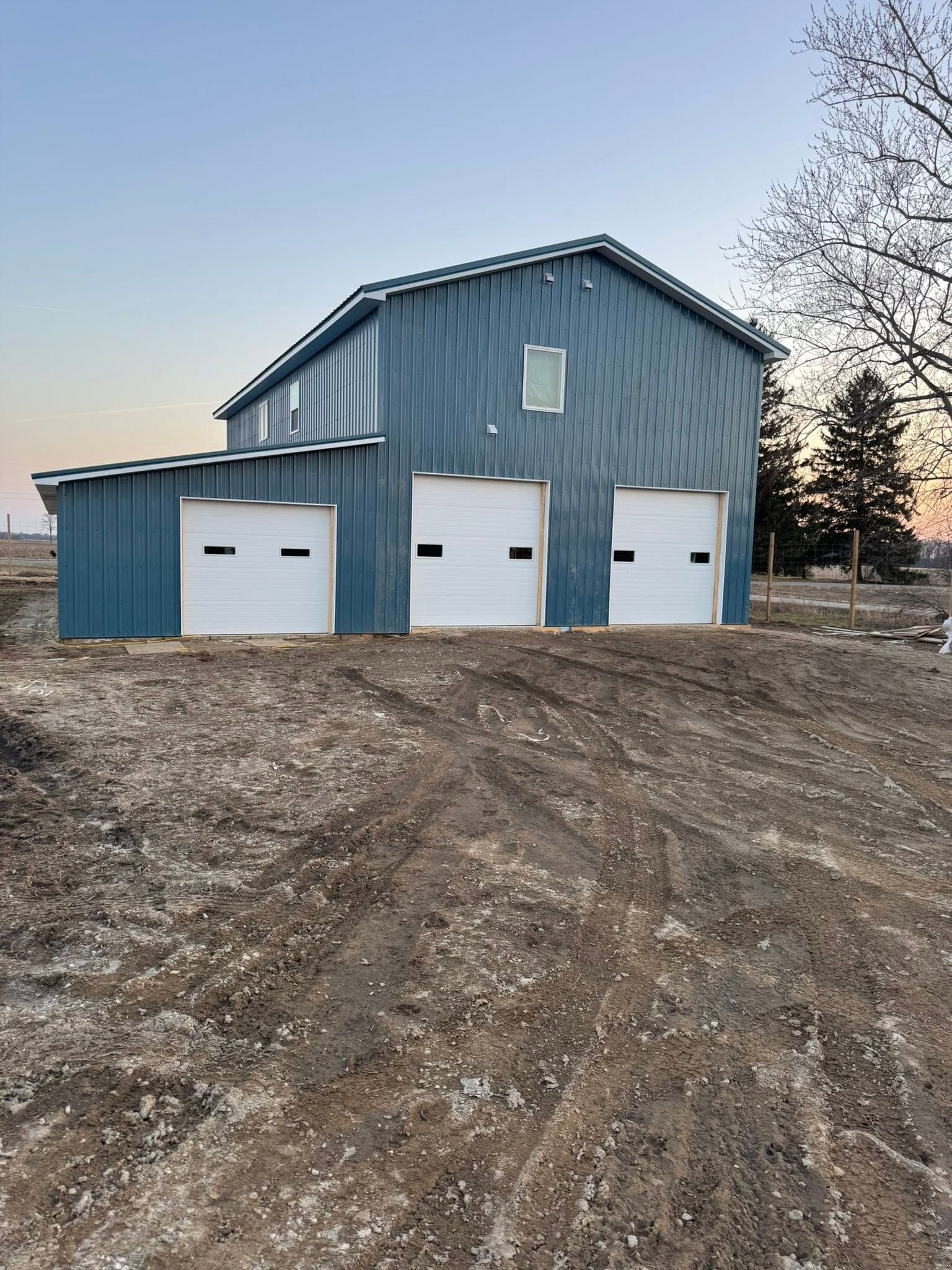 Blue metal garage with three white doors on a dirt lot at dusk.
