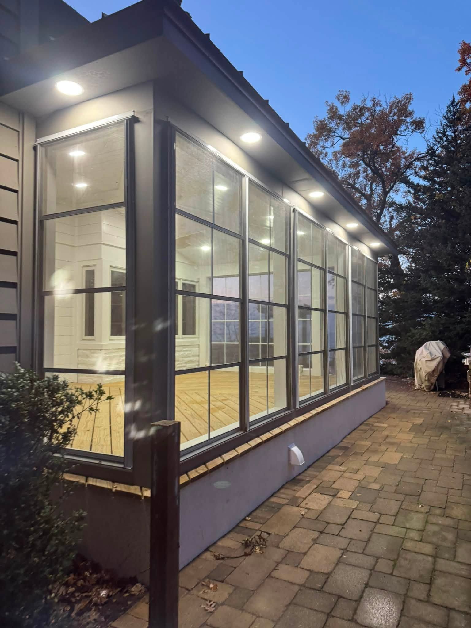 Glass-walled sunroom at dusk beside a brick patio, with warm interior lights and a tree in the background