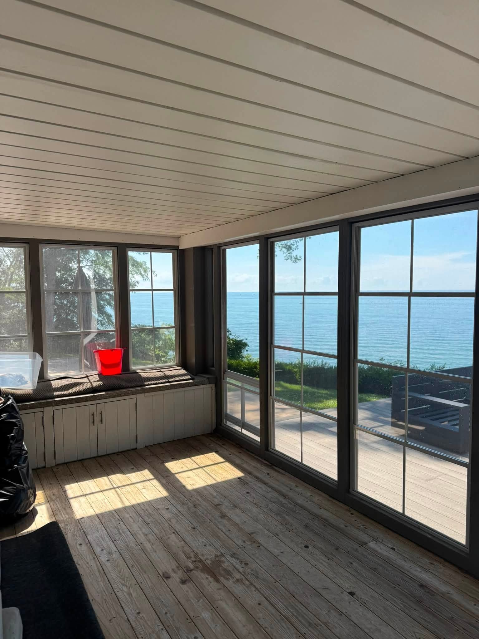 Sunlit room with large windows overlooking the ocean, featuring a red bucket and wooden floorboards.