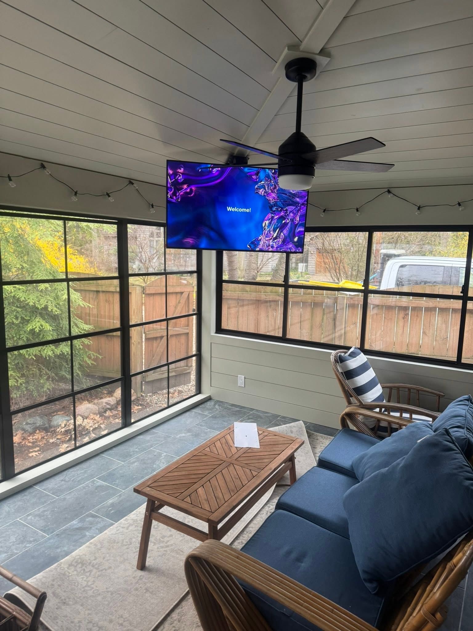 Screened porch with blue sofa, coffee table, hanging TV, and black ceiling fan overlooking a yard.