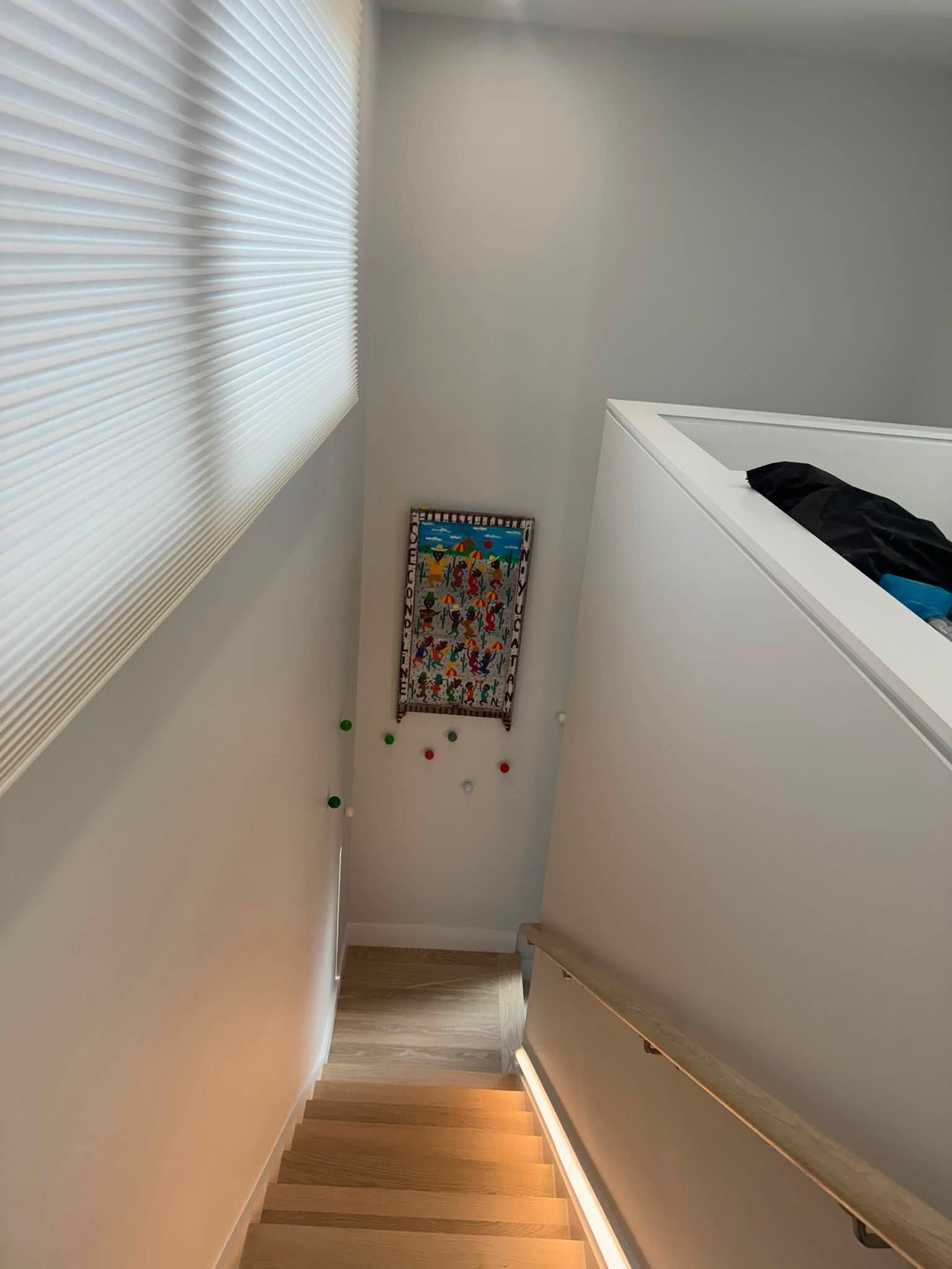 Narrow stairwell with wood steps, white walls, blinds, and a colorful framed print at the bottom landing