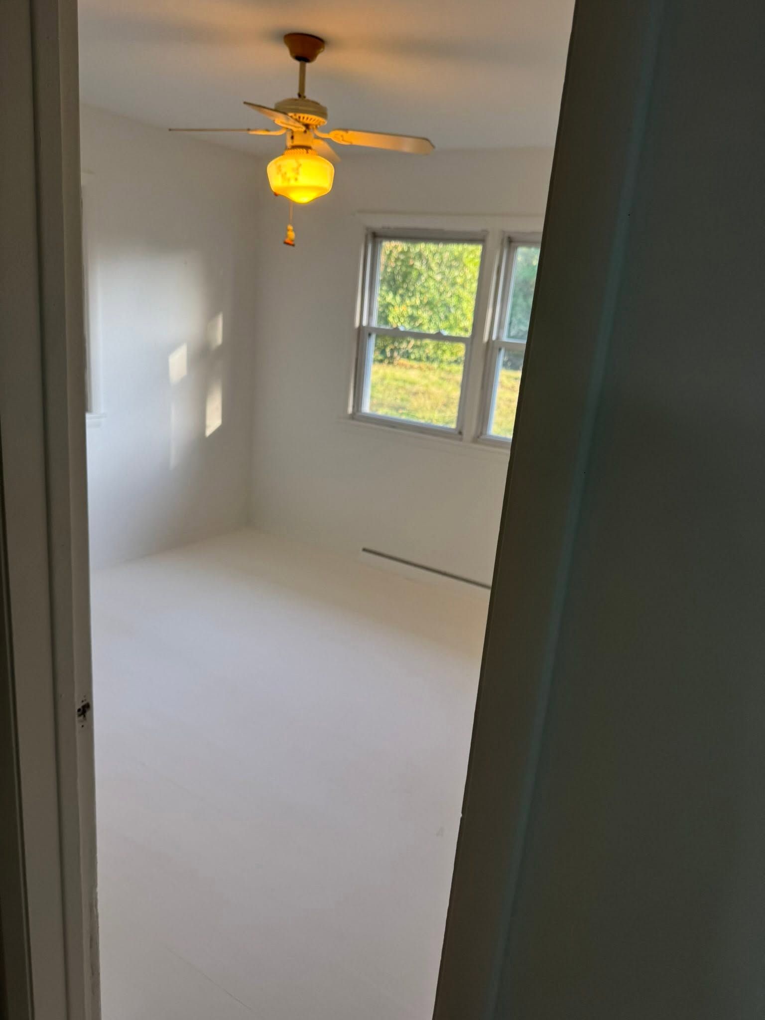 Empty white bedroom with ceiling fan, window, and a hanging light fixture.