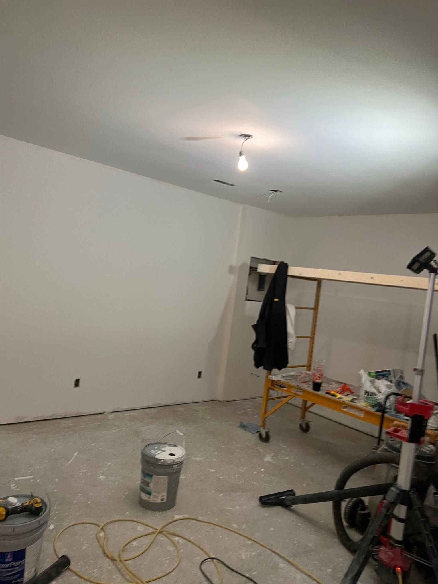 Partially finished room with white walls, ceiling light, ladder, tools, and paint supplies on the floor.