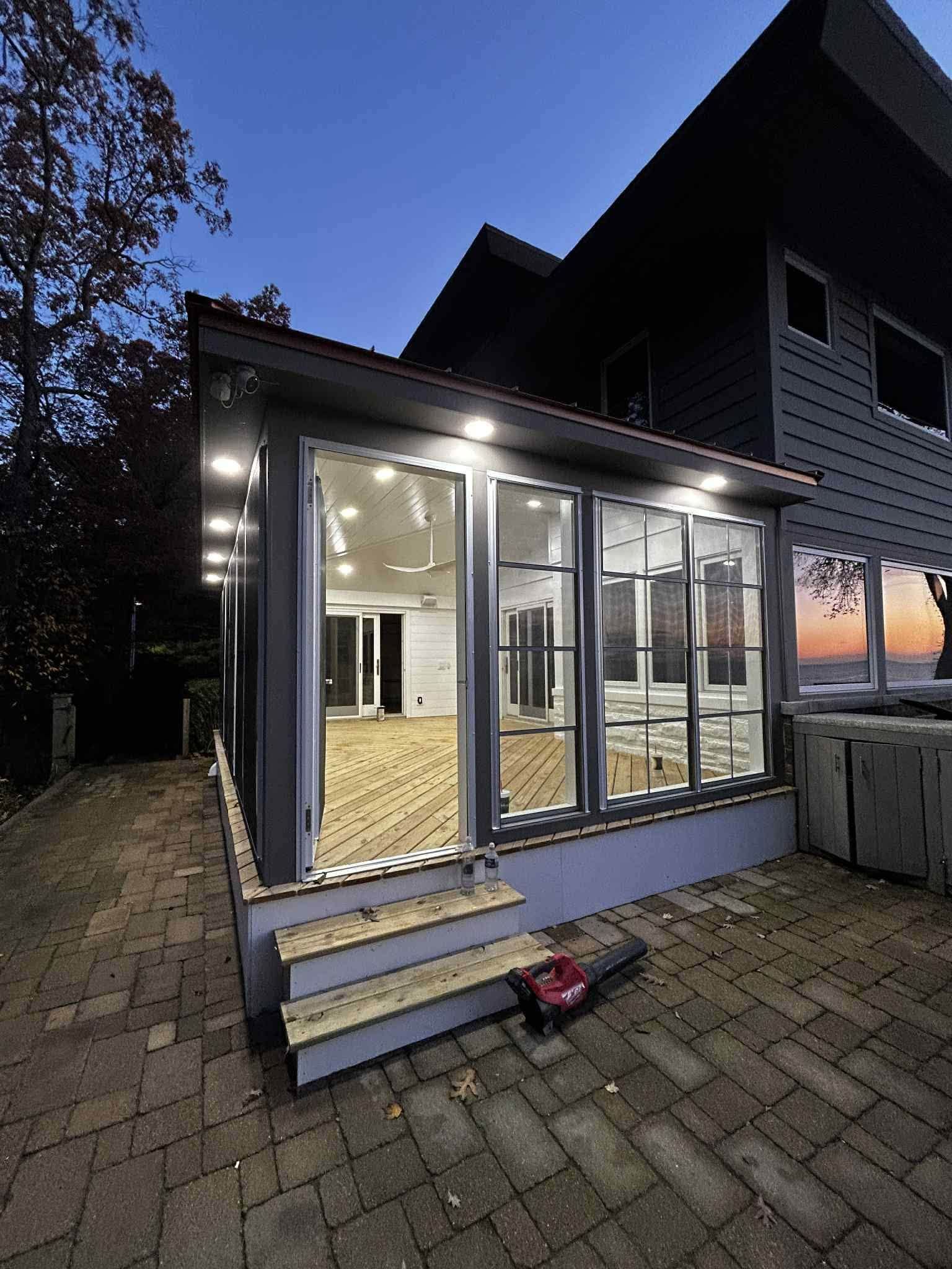 Modern glass-enclosed porch attached to a dark house at dusk, with steps, lit interior, and patio stones.