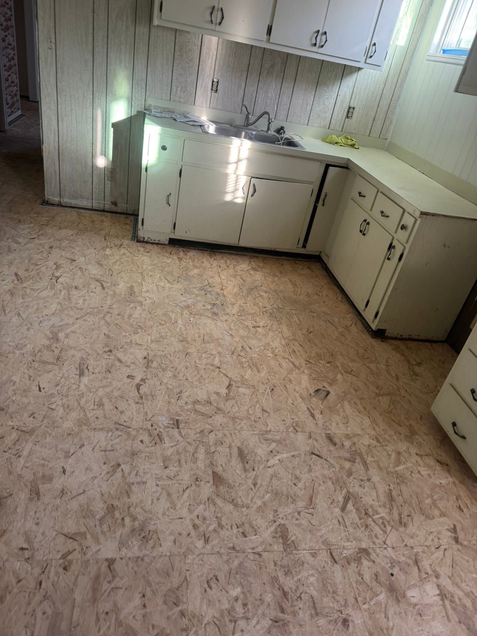 Small kitchen with white cabinets, sink, and speckled beige flooring, showing a worn, empty room.