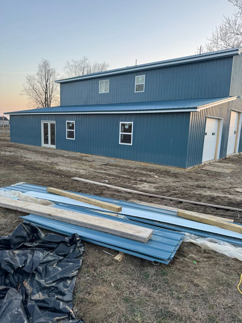 Blue two-story building under construction with metal siding, tarps, and stacked panels in the foreground