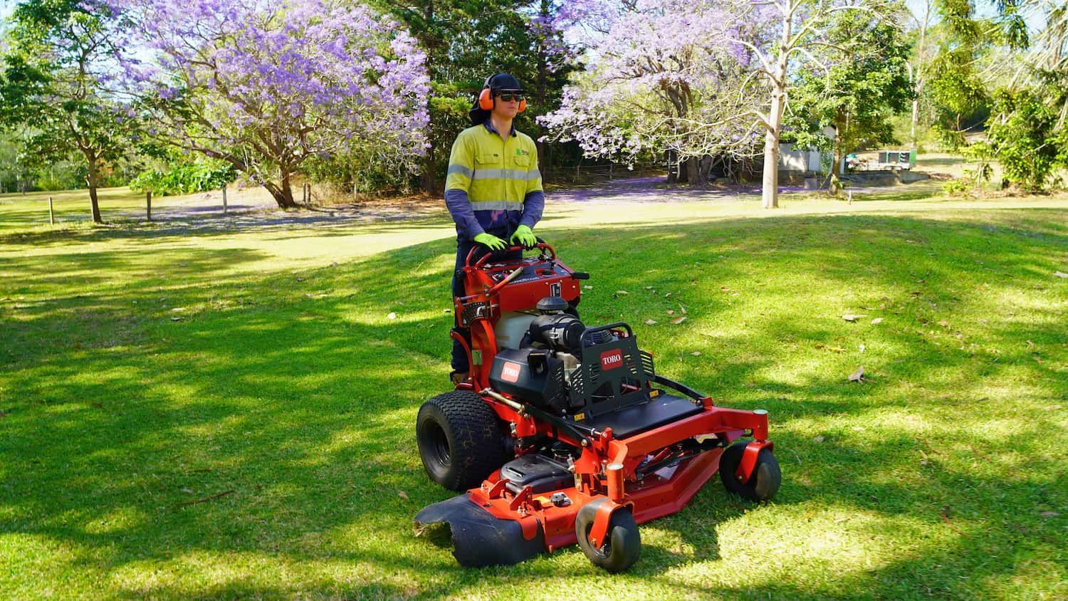 Person operating a red zero-turn mower on a grassy lawn under flowering trees. They wear safety ear protection.