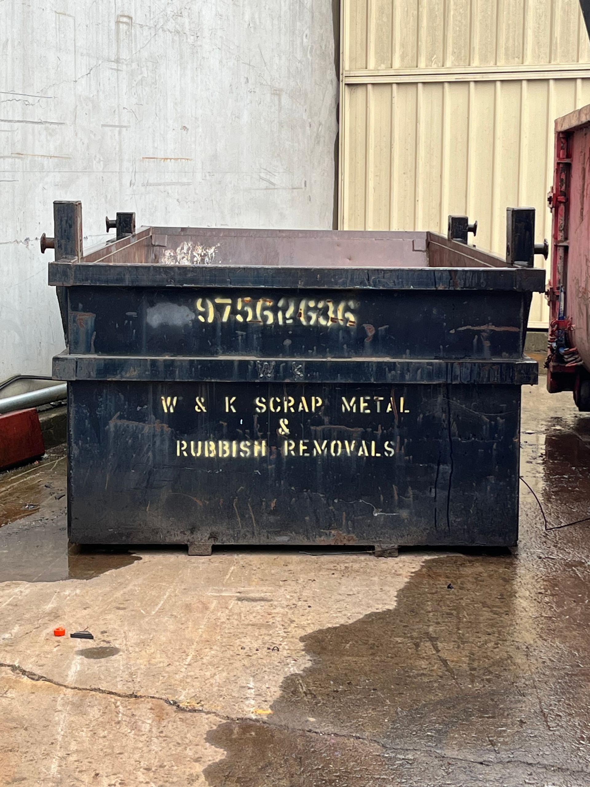 Black metal dumpster with phone number and business name, outdoors on wet pavement.