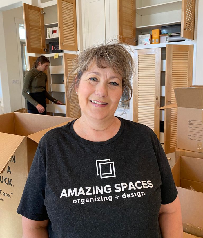 Team member in black “Amazing Spaces organizing + design” T-shirt standing among moving boxes in a room