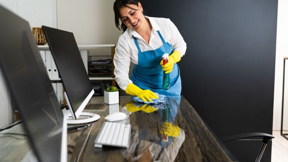 Woman in apron and gloves cleaning an office desk with spray bottle and cloth.