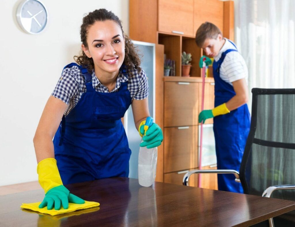 Woman in blue apron smiles while cleaning a wooden table with a cloth; man mops in the background.