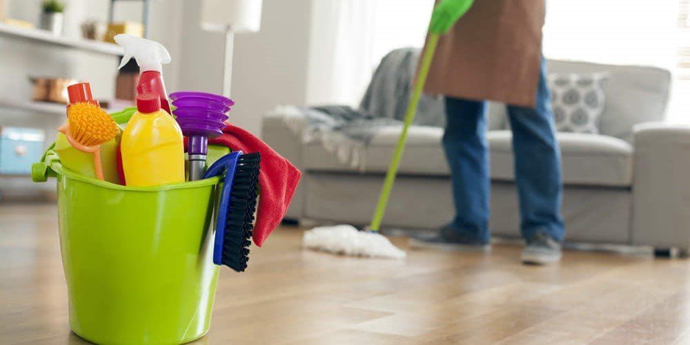 Person mopping a wooden floor, green bucket of cleaning supplies in the foreground, and a couch in the background.
