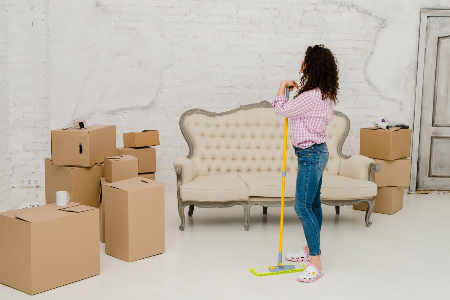 Woman leaning on a mop, surrounded by moving boxes and a sofa, in a bright room.
