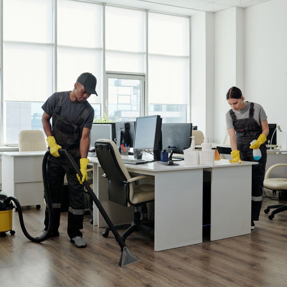 Two people cleaning an office: one vacuums, the other sprays and wipes a desk. They wear gloves and overalls.
