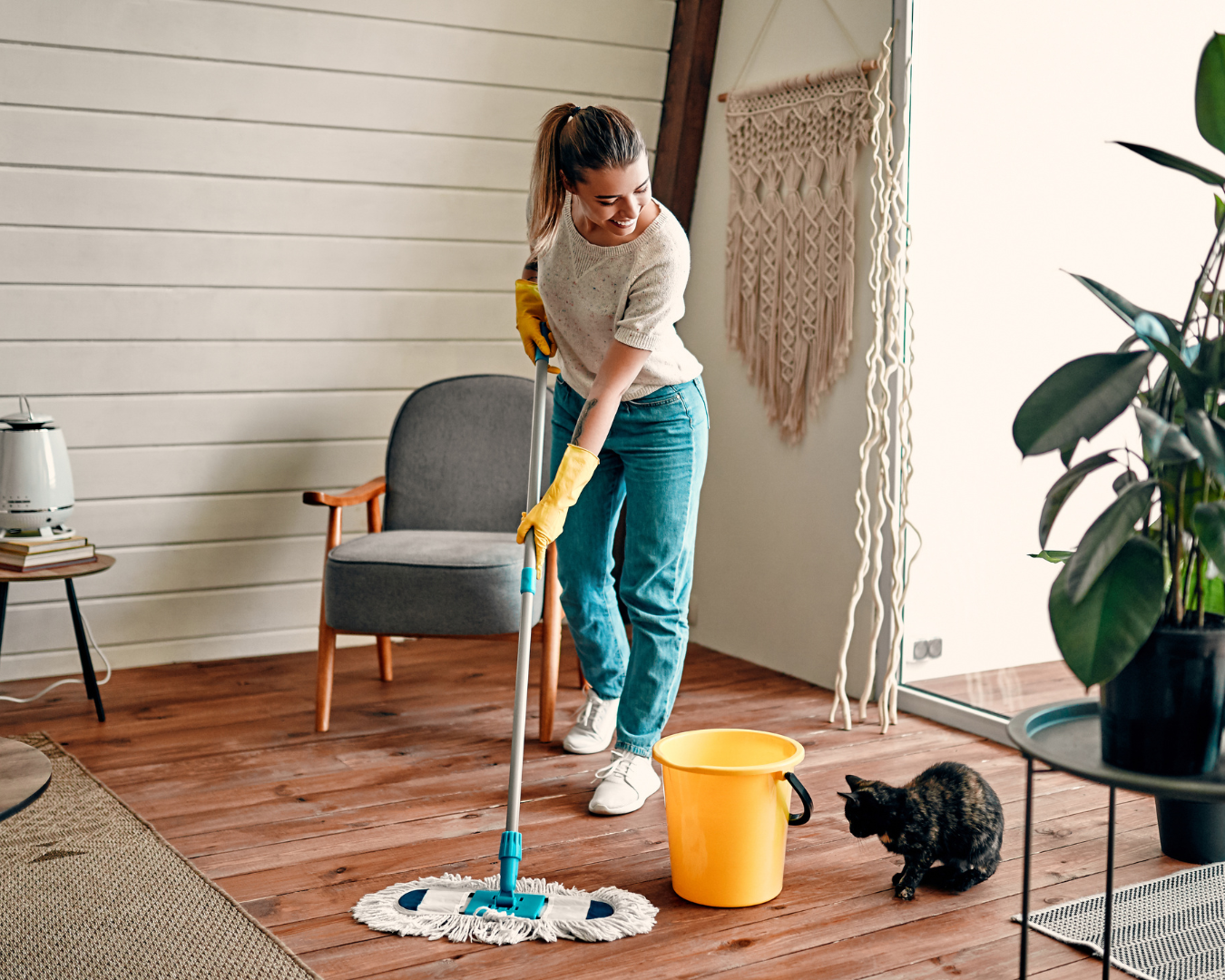 Woman mopping wooden floor with yellow gloves and bucket, cat watching.