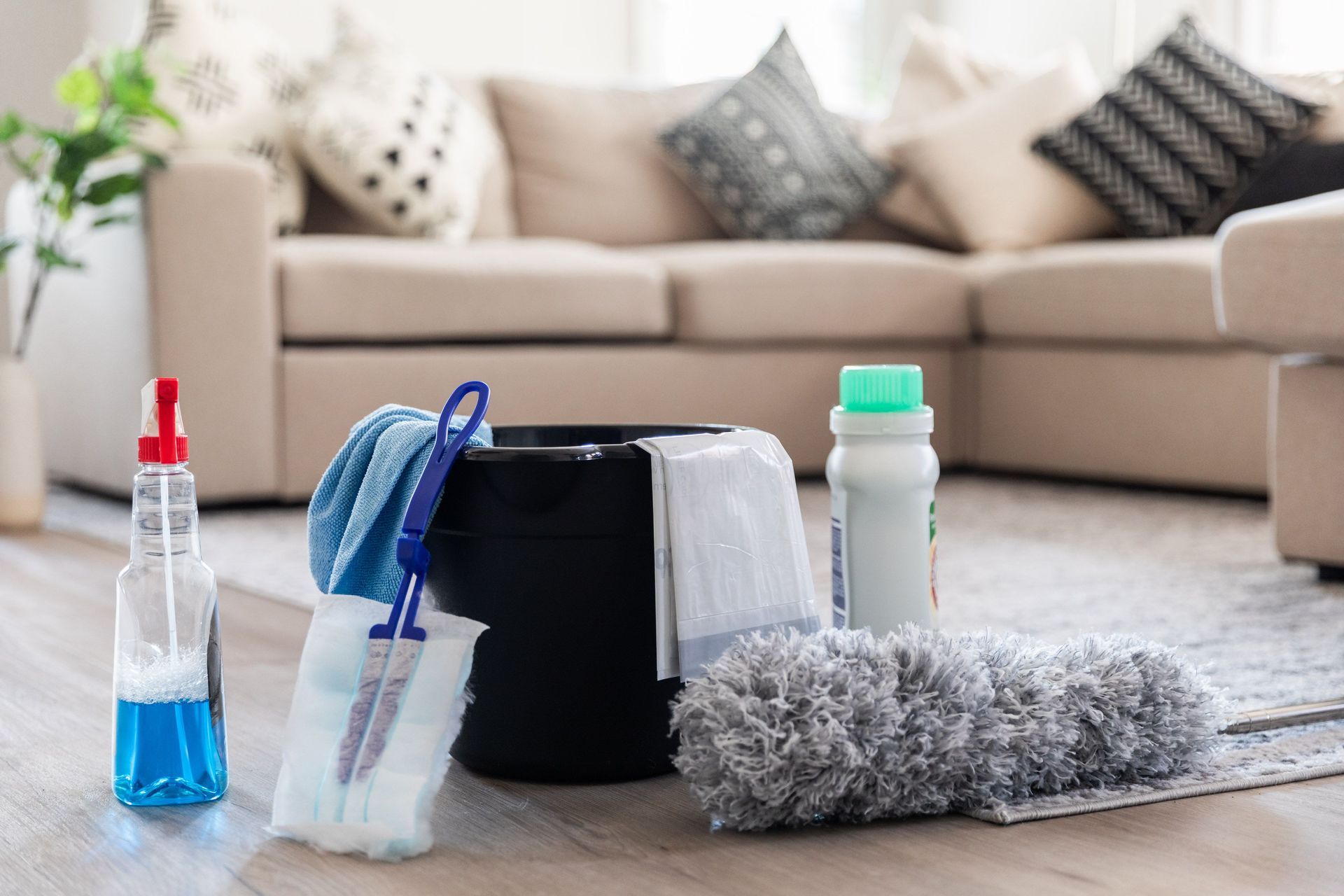 Cleaning supplies (spray bottle, mop, bucket) on a wood floor in front of a beige sectional sofa.