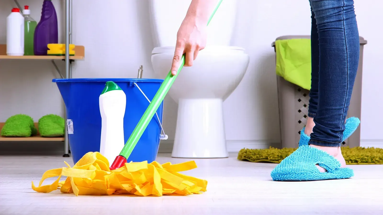 Person mopping a bathroom floor with a yellow mop, blue bucket, and white toilet in the background.