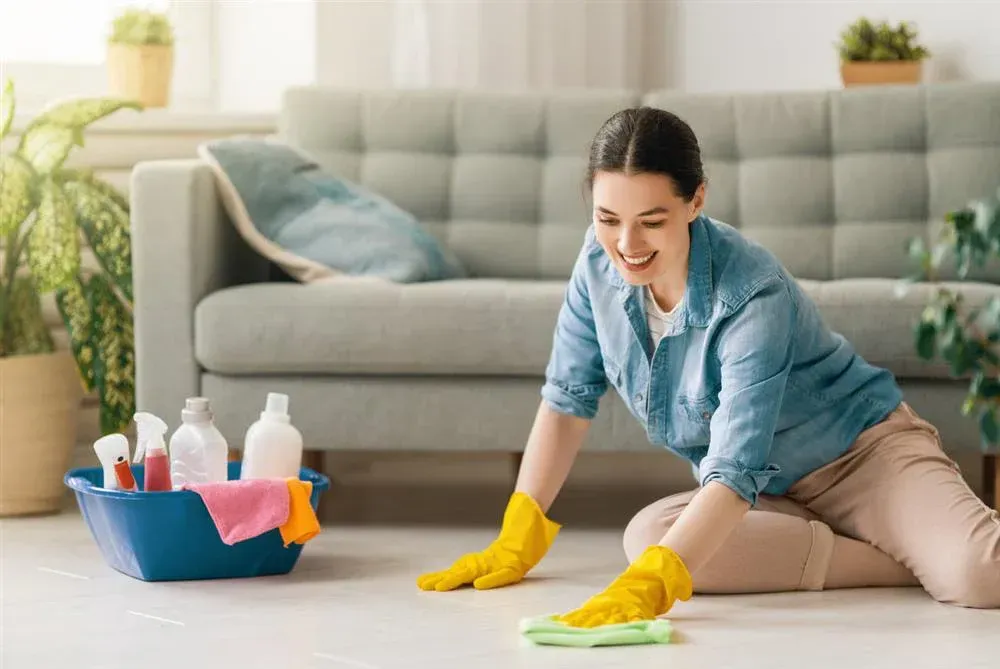 Woman wearing yellow gloves cleaning floor, smiling, in living room with cleaning supplies.