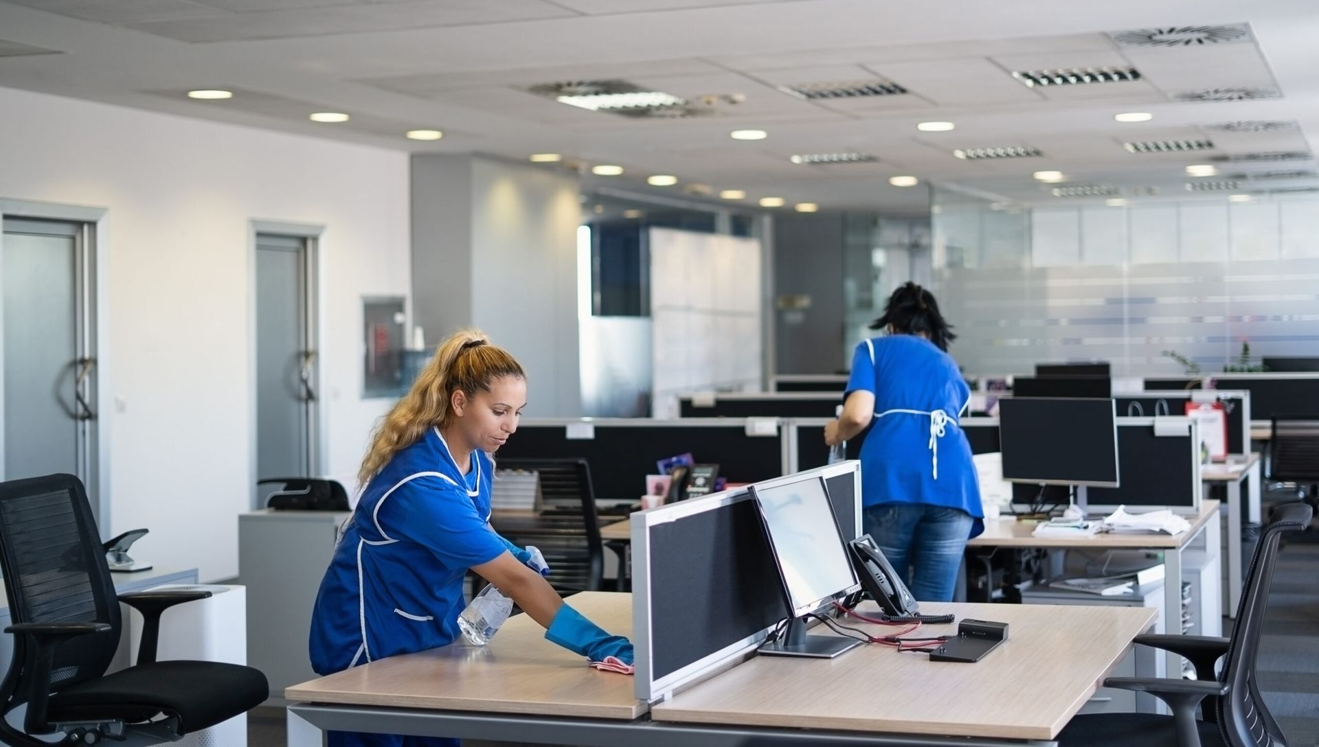 Two office cleaners in blue uniforms sanitizing desks in a modern office.