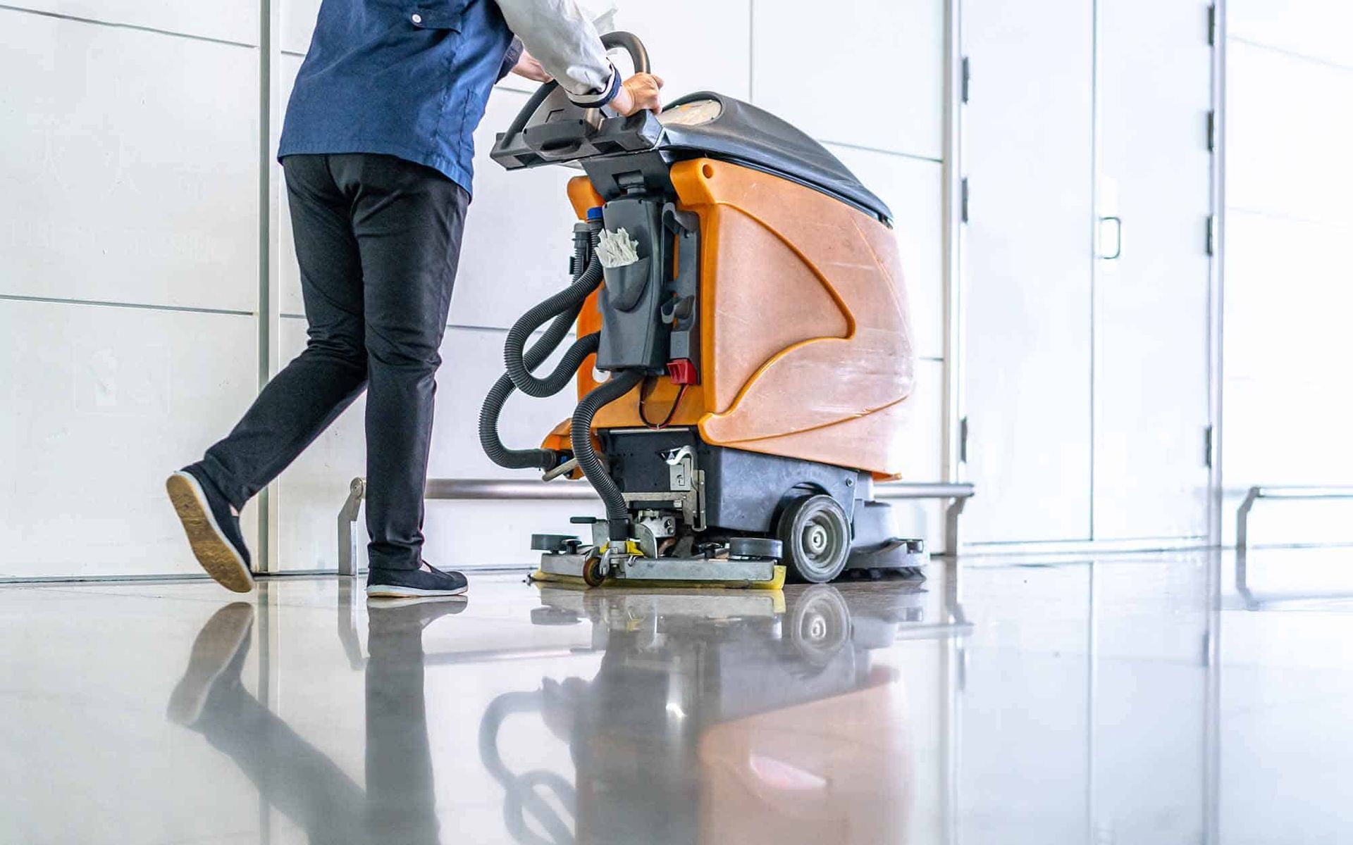 Person operating a floor cleaning machine on a shiny, reflective floor. The machine is orange and black.