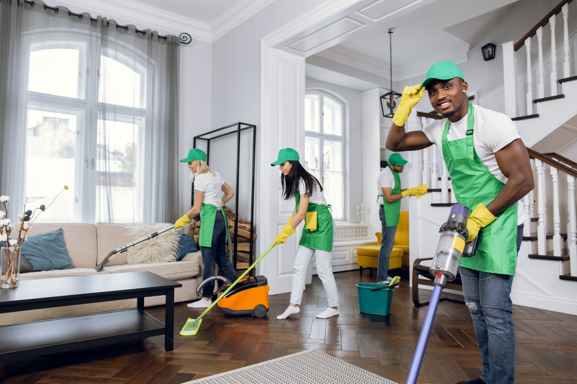 Four cleaning workers in green aprons vacuum and mop a brightly lit living room.
