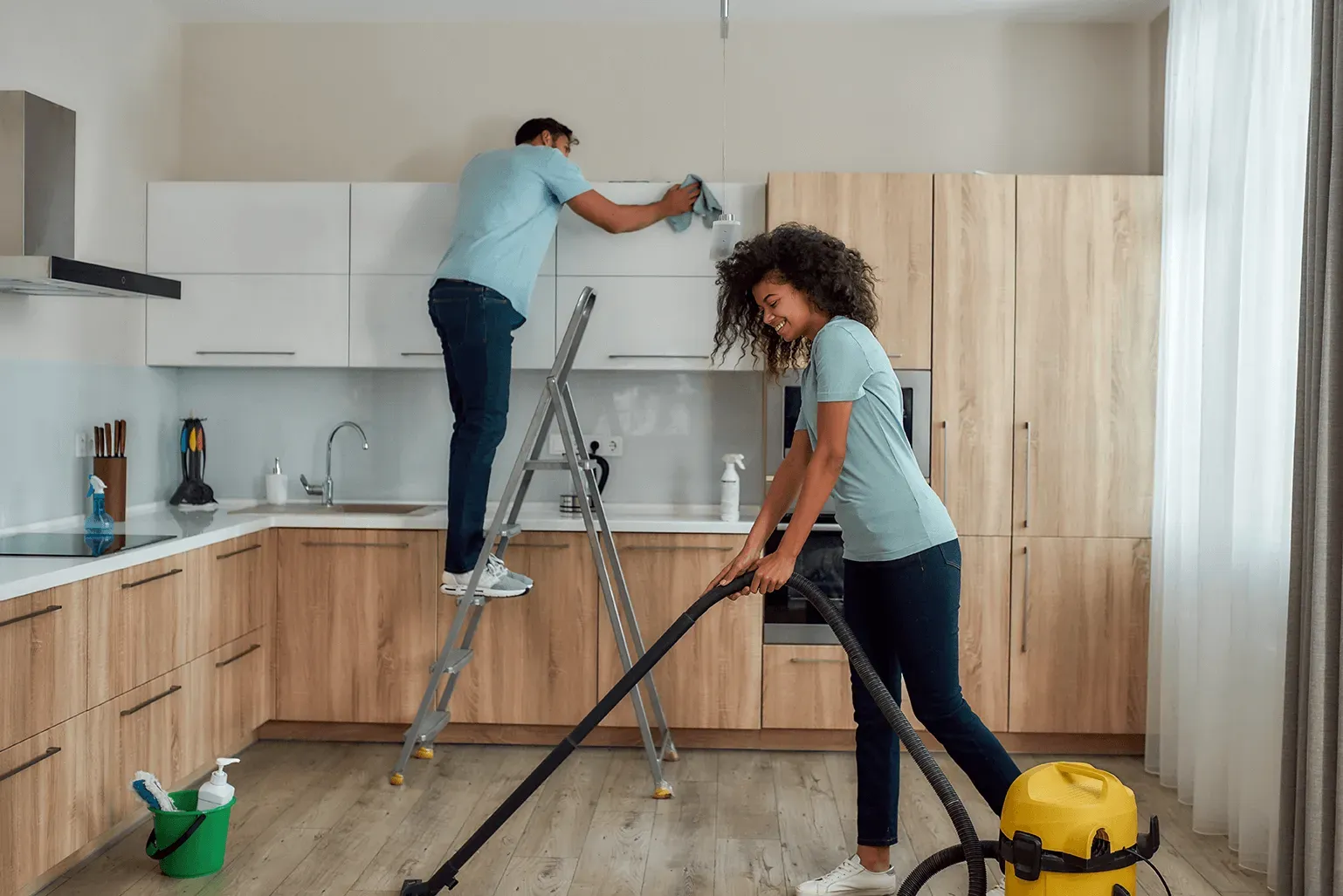 Couple cleaning a kitchen: man on ladder dusting cabinets, woman vacuuming floor. Light wood cabinets, neutral colors.